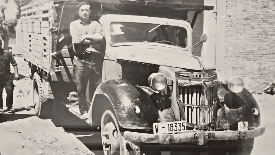 Fotografía antigua en blanco y negro: un hombre con boina, con los brazos cruzados, junto a su camión de carga